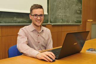 uśmiechnięty Jakub Misiak, student z SKN Cirkula, siedzi przy brązowym biurku. Na nim otwarty laptop. W tle szkolne zielone tablice.