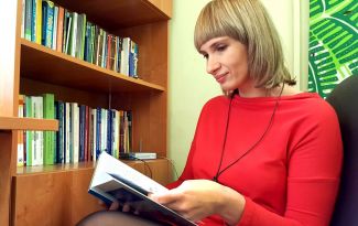 Zdjęcie portretowe: dr hab. Małgorzata Koszewska w czerwonej bluzce siedzi i czyta książkę. W tle regał z książkami. 