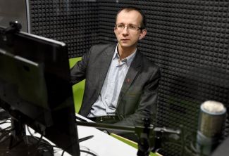 Zdjęcie portretowe: dr inż. Bartłomiej Stasiak siedzi w studiu nagraniowym. Po lewej stronie tył monitora, po prawej fragment mikrofonu.