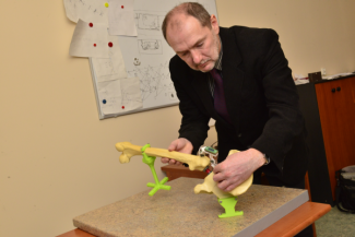 Portrait photo: Prof. Leszek Podsędkowski stands at his desk and assembles a model with yellow and green elements. A board with documents in the background.