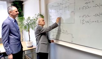 Prof. dr hab. Michał Tadeusiewicz pisze wzór techniczny na białej tablicy. Po lewej stronie stoi dr hab. Stanisław Hałgas, prof. PŁ i przygląda się wzorom.