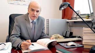 prof. dr hab. Michał Tadeusiewicz w szarym garniturze siedzi przy biurku na który otwarta książka.