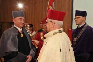 Prof. Stanisław Bielecki, TUL rector, in red gown and white ermine is resting a scepter on the shoulder of Sir Jim McDonald in grey gown and is awarding him the DHC title of TUL. In the background, Dr Eng. Sławomir Hausman wearing a gown.