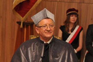 Portrait photo: Prof. Józef Wiesław Modelski in grey gown at the ceremony of awarding the DHC title of TUL. In the background a student from the flag guard with a white-red sash.