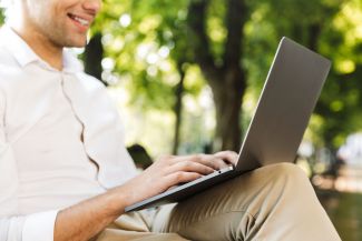 Mężczyzna w białej koszuli i brązowych spodniach trzyma na kolanach laptopa, na którym pisze.