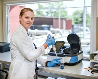 Dr inż. Marta Gmurek w białym fartuchu stoi przy chemicznym stanowisku laboratoryjnym. W ręku trzyma pipetę, którą uzupełnia probówkę.