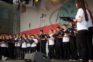 Grupa ok 30 osób w białych t-shirtach i czarnych spodniach, członków Akademickiego Chóru Politechniki Łódzkiej, stoi na scenie podczas występu. W tle kolorowy baner.