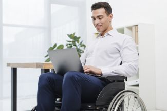 A disabled man in a white shirt is sitting in a wheelchair. He holds a laptop on his lap and works on it.