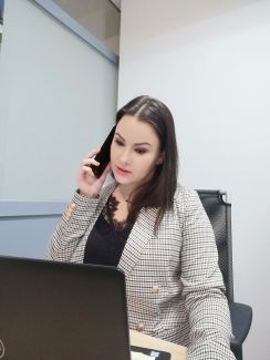 Nina Kozłowska z ciemnej koszuli i marynarce siedzi przy biurku, na którym stoi czarnym laptop. Słuchawkę telefoniczną trzyma przy uchu. W tle biała tablica i ściana.