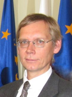 Zdjęcie portretowe: prof. Jacek Jachymski w szarym garniturze i białej koszuli. W tle fragment flagi Unii Europejskiej.