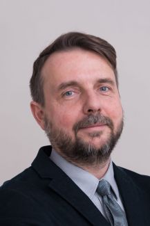 Portrait photo: Prof. Marcin Kamiński wearing a dark jacket, blue shirt and tie a against a grey background.