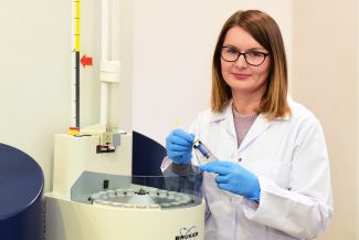 Zdjęcie portretowe: dr inż. B. Pacholczyk-Sienicka w białym fartuchu i niebieskich rękawiczkach stoi przy urządzeniu laboratoryjnym i trzyma w rękach probówkę.