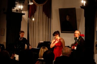 Salka koncertowa w trakcie koncertu cyklu Muzyka na Politechnice. Na scenie, centralnie kobieta w czerwonej sukni, po lewej stronie akompaniatorka przy fortepianie, po prawej prof. J, Krysiński w ciemnym garniturze.