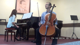 Assoc. Prof. Eng. Bartłomiej Stasiak sits on stage during a concert. A cello in front of him. The accompanist sits at the piano in the background.