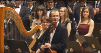 Bartłomiej Stasiak siedzi na scenie podczas przerwy w trakcie koncertu. W tle harfa i orkiestra.