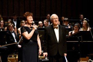 prof. Jan Krysiński w smokingu i Grażyna Sikorska w czarnej sukni stoją na scenie podczas jubileuszu cyklu Muzyka na Politechnice. W tle orkiestra.