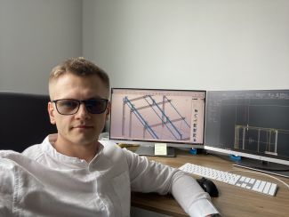 Zdjęcie portretowe: Rafał Bredow w białej koszuli. W tle biurko z białą klawiatura i ekranem na którym widać rysunek techniczny.