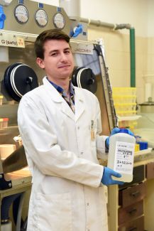 Mgr inż. Andrzej Gąsiorowski stoi frontem w białym fartuchy w laboratorium i w rękach trzyma przeźroczysty pojemnik z nieczytelną etykietą. W tle aparatura chemiczna.