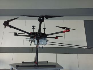 Czarny dron na statywie postawiony w laboratorium, na szarym stole. W centralnym miejscu dron ma podpiętą białą skrzynkę z aparaturą pomiarową.