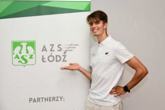 Kajetan Duszyński, doktorant z PŁ w białej polówce stoi na tle biało-zielonej ścianki AZS Łódź i wskazuje na logotyp organizacji.