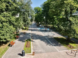 Top photo of the woonerf on Stefanowski Street. On the sides, tall green trees, in the middle, on the left, pavements and benches, on the right, a street of grey cobbles.
