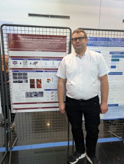 Dr inż. Dominik Knozowski w okularach, białej koszuli z krótkim rękawem i czarnych spodniach stoi obok swojego plakatu naukowego, zawieszonego na metalowym stojaku. Plakat ma tytuł „Plasma-Deposited Cobalt Oxide Thin Films: Nanostructure Engineering for Advanced Electrochemical Catalysis” i logo Politechniki Łódzkiej. 