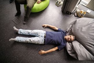 Mężczyzna w granatowej koszulce i jasnych dżinsach leży na podłodze z rozłożonymi rękami, z głową opartą o szarą pufę, podczas ćwiczenia relaksacyjnego. Zdjęcie zrobiono z góry