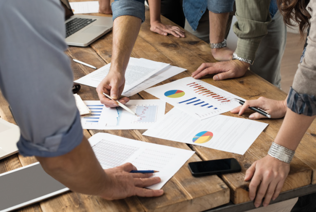 Drewniany st&oacute;ł, na nim kilka kartek w kolorowymi wykresami.