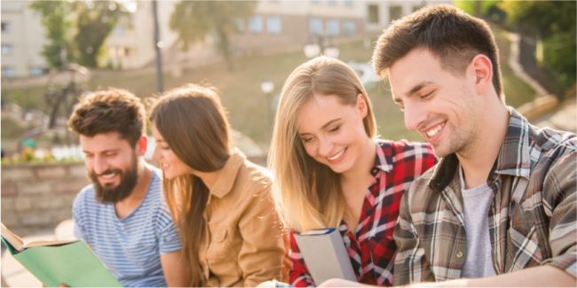 Czworo uśmiechniętych studentów (dwie studentki w środku) przeglądają materiały informacyjne. W tle park.