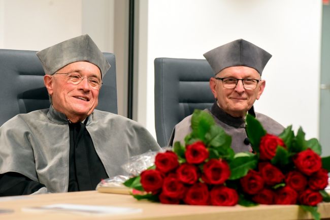 Prof. Kalus Mullen i prof. Jacek Ulański siedzą w togach przy drewnianym stole prezydialnym. Przed nimi leżą bukiety czerwonych róż.