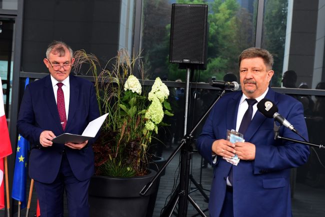 Marszałek Województwa Łódzkiego stoi przy mikrofonie. W tle prof. Krzysztof Jóźwik i ciemna szyba budynku Alchemium.