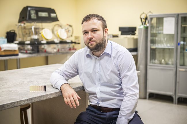 Dr inż. Michał Piotrowski w jasnoniebieskiej koszuli, siedzi w laboratorium i opiera rękę o blat stołu. Obok niego stoi rząd kuwet z próbkami w odcieniach od jasnożółtego do ciemnofioletowego.