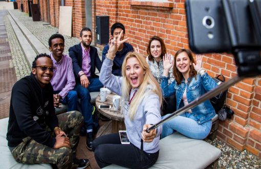 Seven, smiling international students sit in the city centre. A girl holds an unfolded selfie stick and phone and takes a picture.