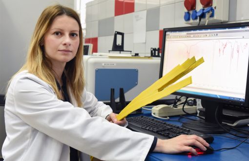 Portrait photo: Dr Eng. Anna Masek in a white coat is sitting at her desk in right profile. In her hand she is holding yellow stripes. On the right, a computer screen with data. In the background, a laboratory. 