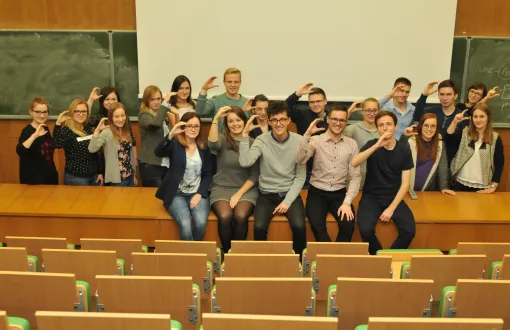 Zdjęcie portretowe: grupa ok 30 studentów z SKN Cirkula  w sali wykładowej na katedrze. Prawe dłonie mają ułożone w literę C. Z tyłu biała i zielone, szkolne tablice.