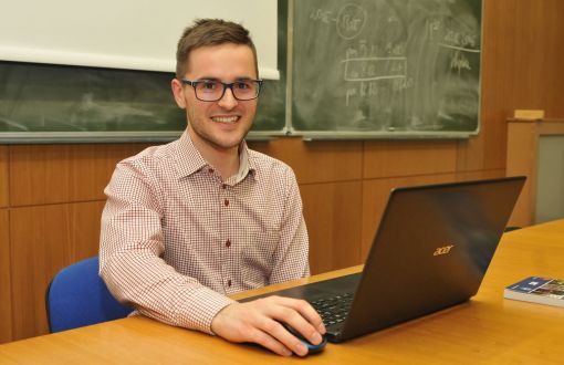 uśmiechnięty Jakub Misiak, student z SKN Cirkula, siedzi przy brązowym biurku. Na nim otwarty laptop. W tle szkolne zielone tablice.