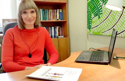 Zdjęcie portretowa: dr hab. Małgorzata Koszewska w czerwonej bluzce siedzi przy biurku. NA nim dokumenty i komputer. W tle zielona grafika i regał z książkami.