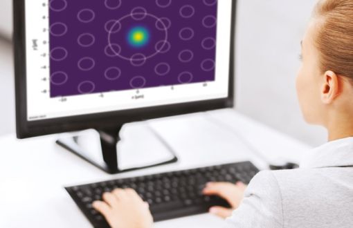 Ekran komputera na którym widać nierównomiernie rozłożone kółka na fioletowym tle. Widoczny także fragment kobiety w białym fartuchu pracującej na komputerze.