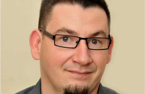 Zdjęcie portretowe: prof. Przemysław Perlikowski w szarej koszuli na jasnym tle.