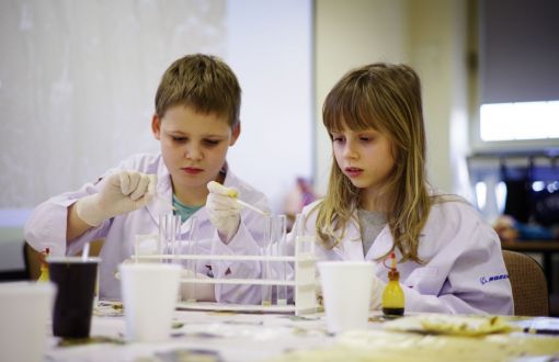 Chłopiec i dziewczynka w białych fartuchach przy stole laboratoryjnym.
