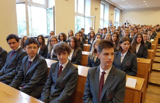 Uczniowie Liceum Politechniki Łódzkiej w szarych marynarkach i bordowych krawatach siedzą w auli przy drewnianych ławach.