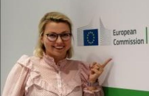 Zdjęcie portretowe: uśmiechnięta Kaja Kantorska na tle jasnej ściany z logiem European Comission, które wskazuje palcem wskazującym.