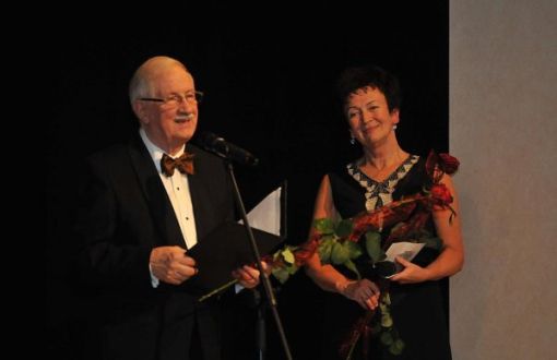 Prof. Jan Krysiński w smokingu i Grażyna Sikorska w czarnej sukni na scenie stoją przy mikrofonie na czarnym tle.