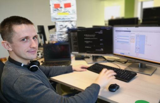 Łukasz Wiśniewski, absolwent PŁ, siedzi przy biurku na którym stoją 2 czarne monitory i klawiatura.