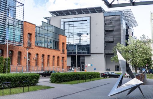 Kampus A Politechniki Łódzkiej: po lewej stronie fragment ceglanego, dwupiętrowego bydynku IFE ze szklanymi elementami. W tle nowoczesna, wysoka, szara bryła - budynek LabFactor. Po prawej stronie metalowa, abstrakcyjna rzeźba.