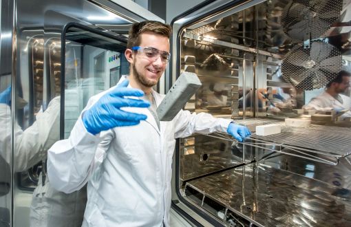 Mateusz Łukasik w białym fartuchu i niebieskich rękawiczkach przy urządzeniu laboratoryjnym. W tle jego odbicia z różnej perspektywy.