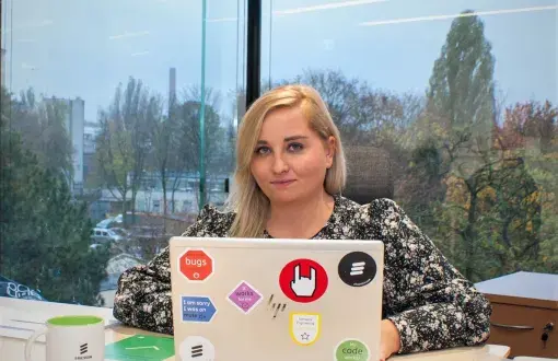 Sylwia Kobus, absolwentka PŁ, przy biurku i laptopie. W tle przeszklone okno z widokiem na park.