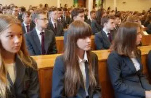 Uczniowie Liceum Politechniki Łódzkiej w szarych marynarkach i bordowych apaszkach (uczennice) i krawatach (uczniowie) siedzą w auli przy drewnianych ławach.