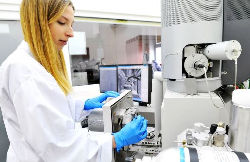 Po lewej stronie stoi młoda kobieta w białym fartuchu i niebieskich rękawiczkach i pracuje przy dużym, białym urządzeniu laboratoryjnym.