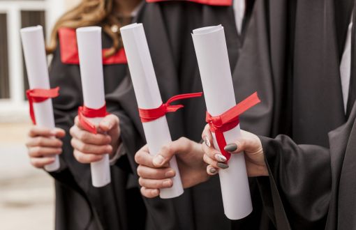 Four hands in robes holding white diplomas rolled up and tied with red ribbon.